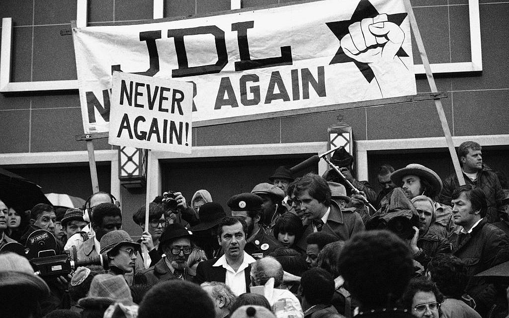 A black and white photo of protestors in a dense crowd. Over them hangs a banner that reads "JDL NEVER AGAIN" and a second banner that reads "NEVER AGAIN!" On one banner is a star of david with a fist held infront of it as a symbol.
