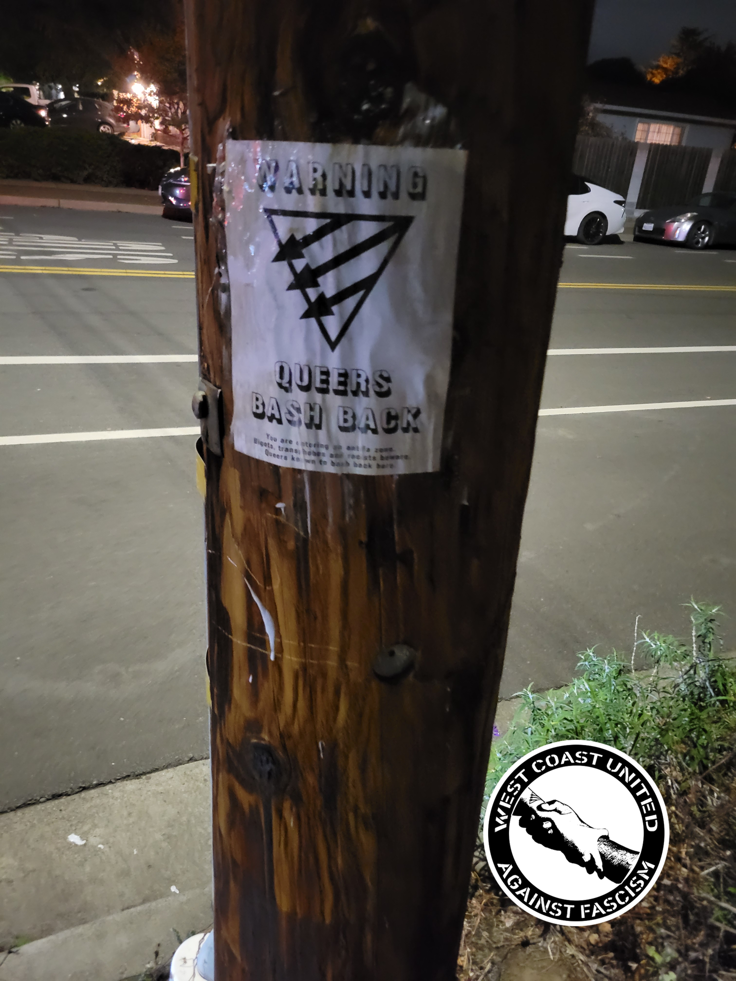 A wooden post has a flyer attached to it. It reads "Warning, Queers Bash Back" with a Queer Front symbol on it, a triangle with three arrows.