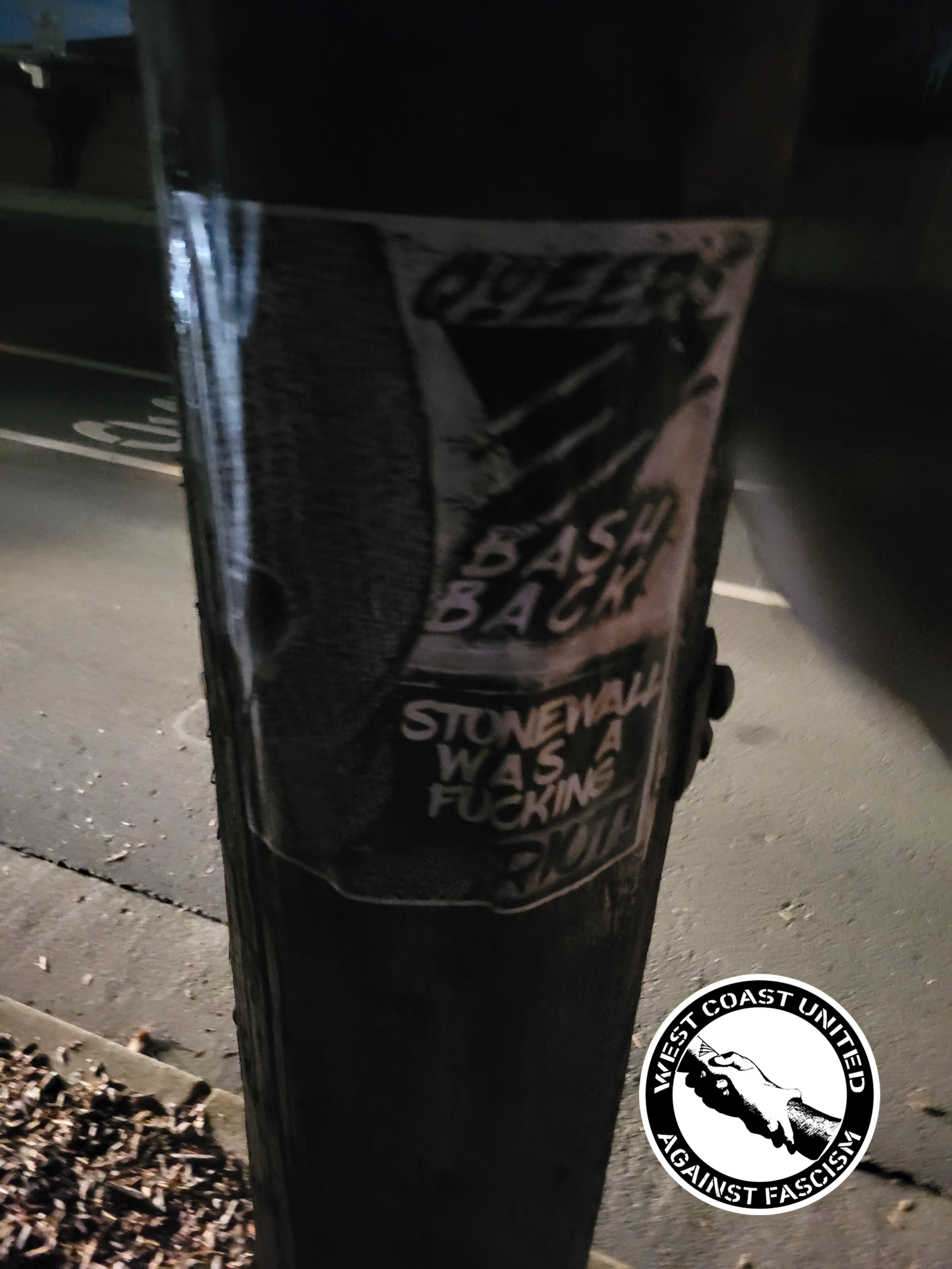 A wooden post has a flyer attached to it. It shows a ski-mask on half the page. On the other half, it reads "Queers bash back" with a traingle with three bats going across it. The bats have spikes. Below it reads "Stonewall was a fucking riot!"