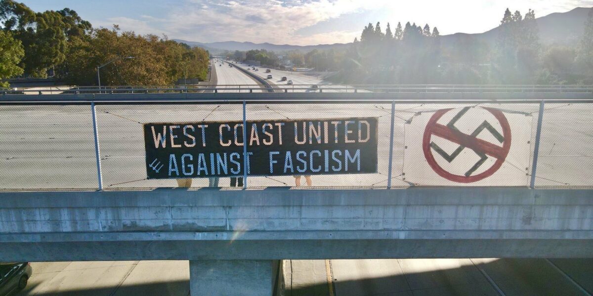 A black banner with white text tied to a freeway overpass that reads "West Coast United Against Fascism" with three arrows in the corner. A canvas is draped next to it with a crossed out swastika. Cars drive underneeth.