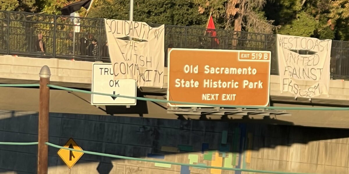 Two large canvas sheets hang over the side of an active freeway. One reads "Solidarity with the jewish community." The other reads "West coast united against fascism." There are red and black flags waving on the overpass, road flares to the side and barely visible bodies in black clothes behind the fence. The traffic sign reads "Exit 518 Old Sacramento State Historic Park Next Exit."