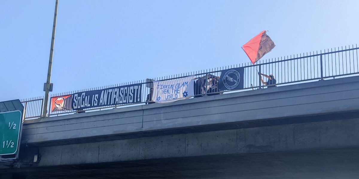 Banners hang off the 405 Freeway that read "SoCal is Antifascist" "Tikkun Olam!" heal the world with a Star of David on each side. A third canvas is the West Coast United Against Fascism with two hands gripping each others arms. To the side is someone waving a red and black flag from the top to the background of a clear blue sky.