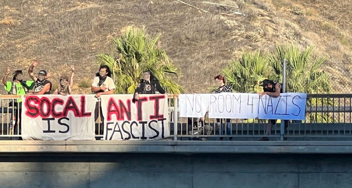 A group of studded jacket punks drop banners off the side of a freeway that read "SoCal is Antifascist" in black and red. A second banner reads "No room 4 Nazis."