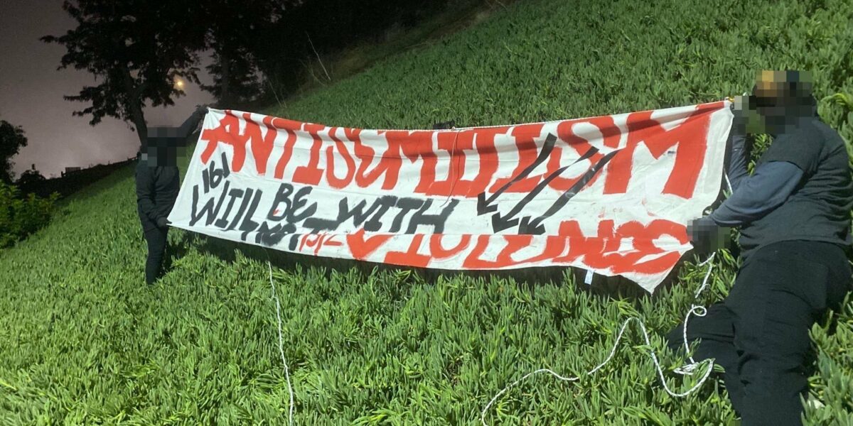 Two antifascists in black clothing drape a white banner with red and black text that reads "Antisemitism will be met with violence." with three arrows down the side.