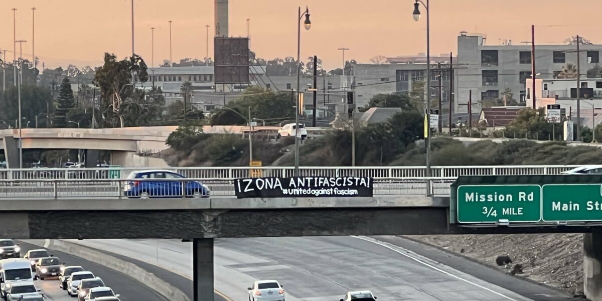 A black banner stretched over the side of an overpass atop a busy freeway with cars passing under. The banner reads "iZona Antifascista! #unitedagainstfascism"