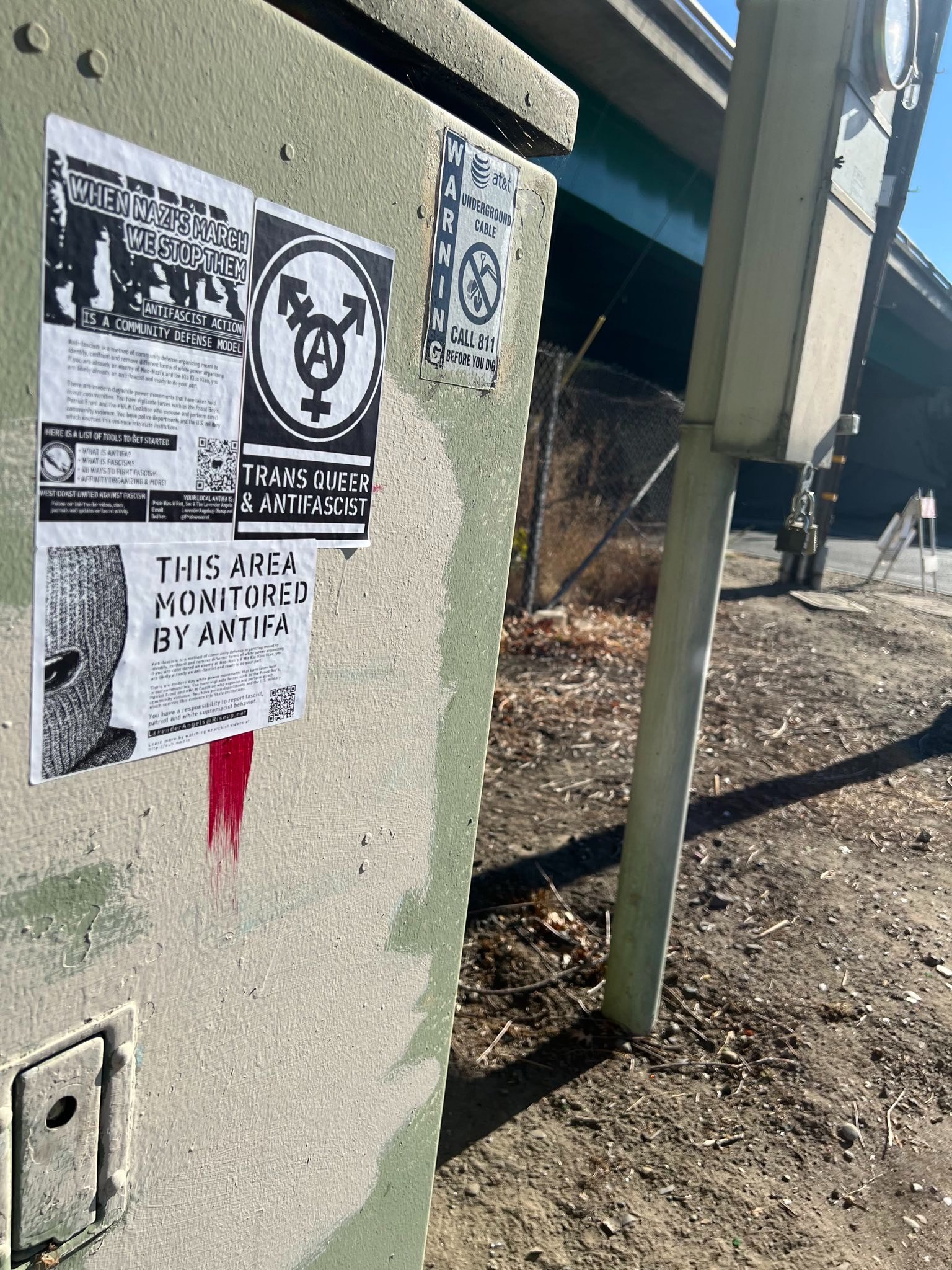 A green electric box with a white supremacist symbol on it has been covered up with antifascist stickers under a nearby overpass.