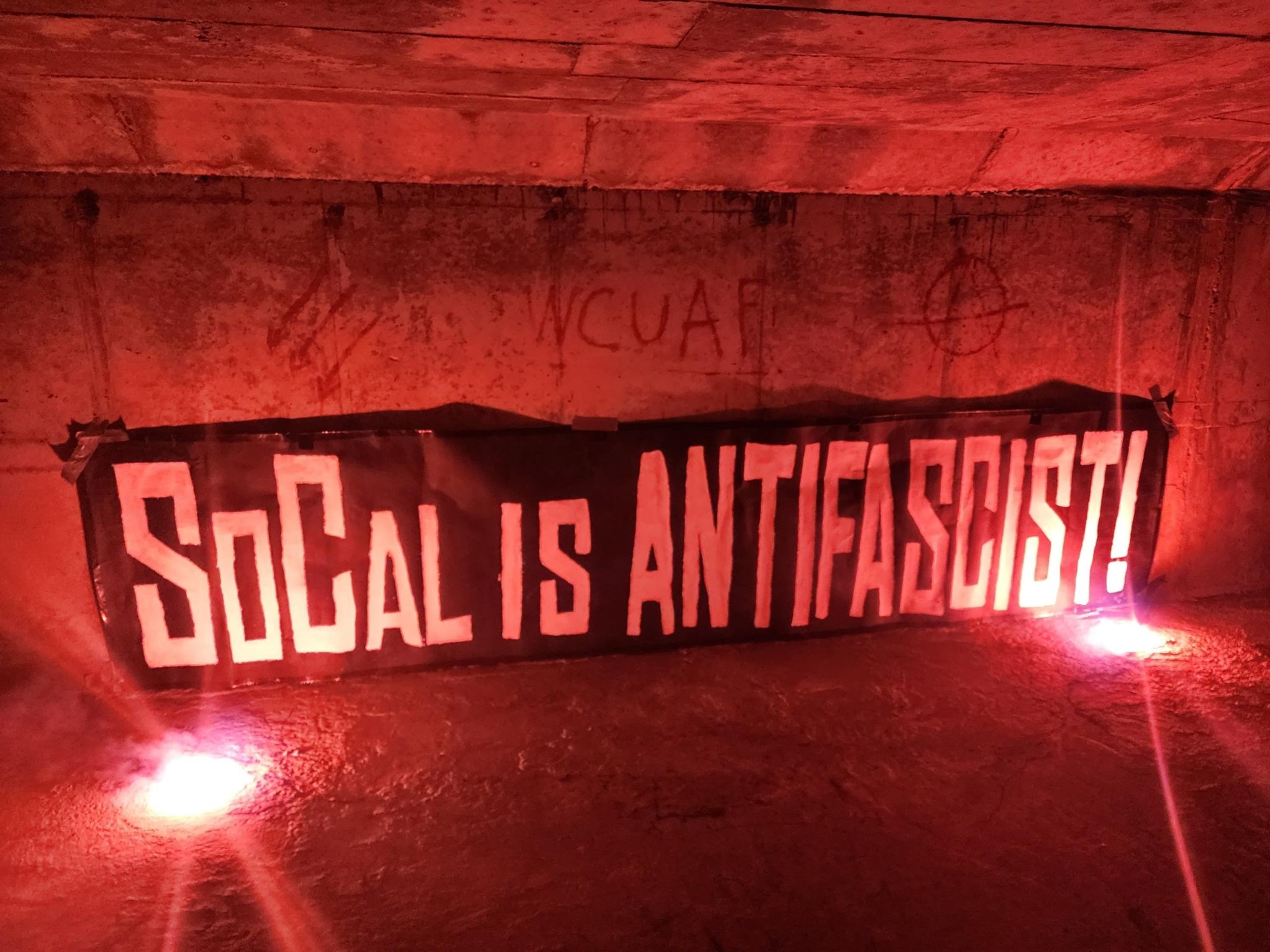 A black banner with white text that reads "SoCal is ANTIFASCIST!" with road flares on either side. Its placed against what appears to be a concrete wall in a dark tunnel. The wall has three arrows, the letters W.C.U.A.F. and an anarchist circle-a behind it.
