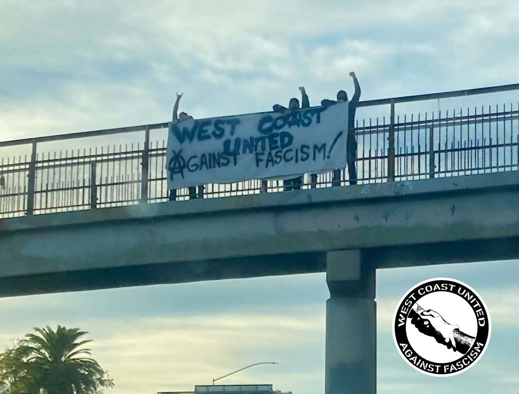 Three people stand on a bridge overpass with the sky behind them. Their fists are up. Infront of them hangs a sign that reads, "West Coast United Against Fascism!" The "a" in against is a circle A, used commonly to symbolize Anarchism.