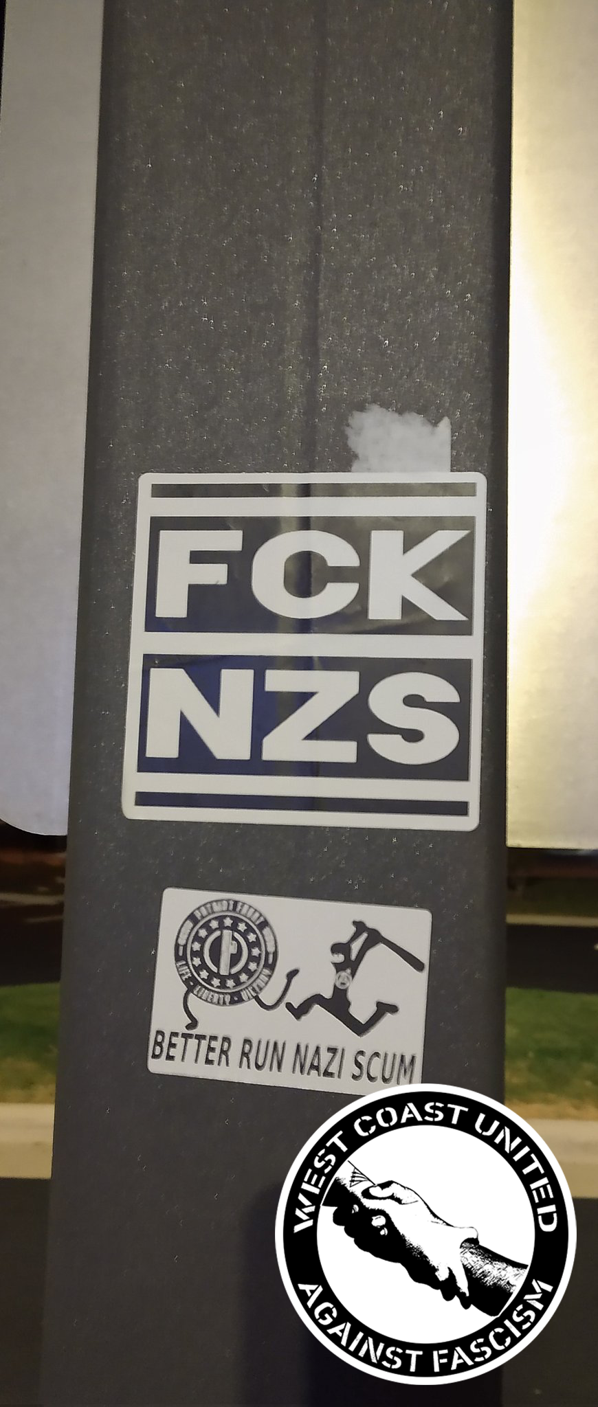 Two images adhear to a metal post. One reads "FCK NZS" in black and white. The one below it shows a picture of the Patriot Front symbol having legs, running away from a stick figure in all black, carrying a bat and wearing a balaclava with an anarchist "circle a" symbol on it. Below reads, "Better Run Nazi Scum"