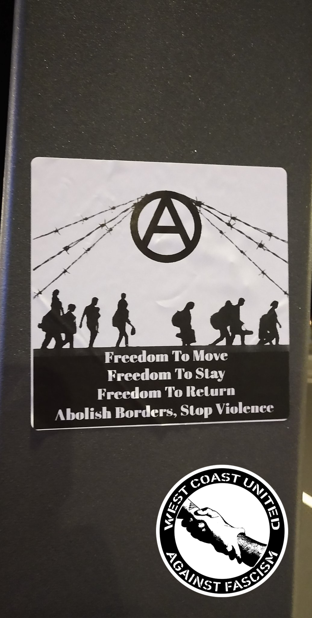 A square sticker is adheared to a metal post. There is a horizon line between black and white where people carrying bags and children are walking from left to right. There is barbed wire going across that is being lifted up by the symol of a "circle a", the common symbol for anarchism. Below the people crossing the barbed wire reads "Freedom to move, Freedom to Stay, Freedom to Return. Abolish borders, stop violence."