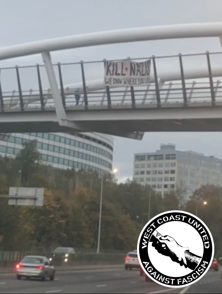 Above a street with heavy traffic is an enourmas bridge of peculiar design. Traffic roars below with trees and city buildings in the background. The bridge has tall, tall fencing and large, dome-like support links. On the fence, a large banner hangs. It reads "Kill Nazis. We Know Where You Live."