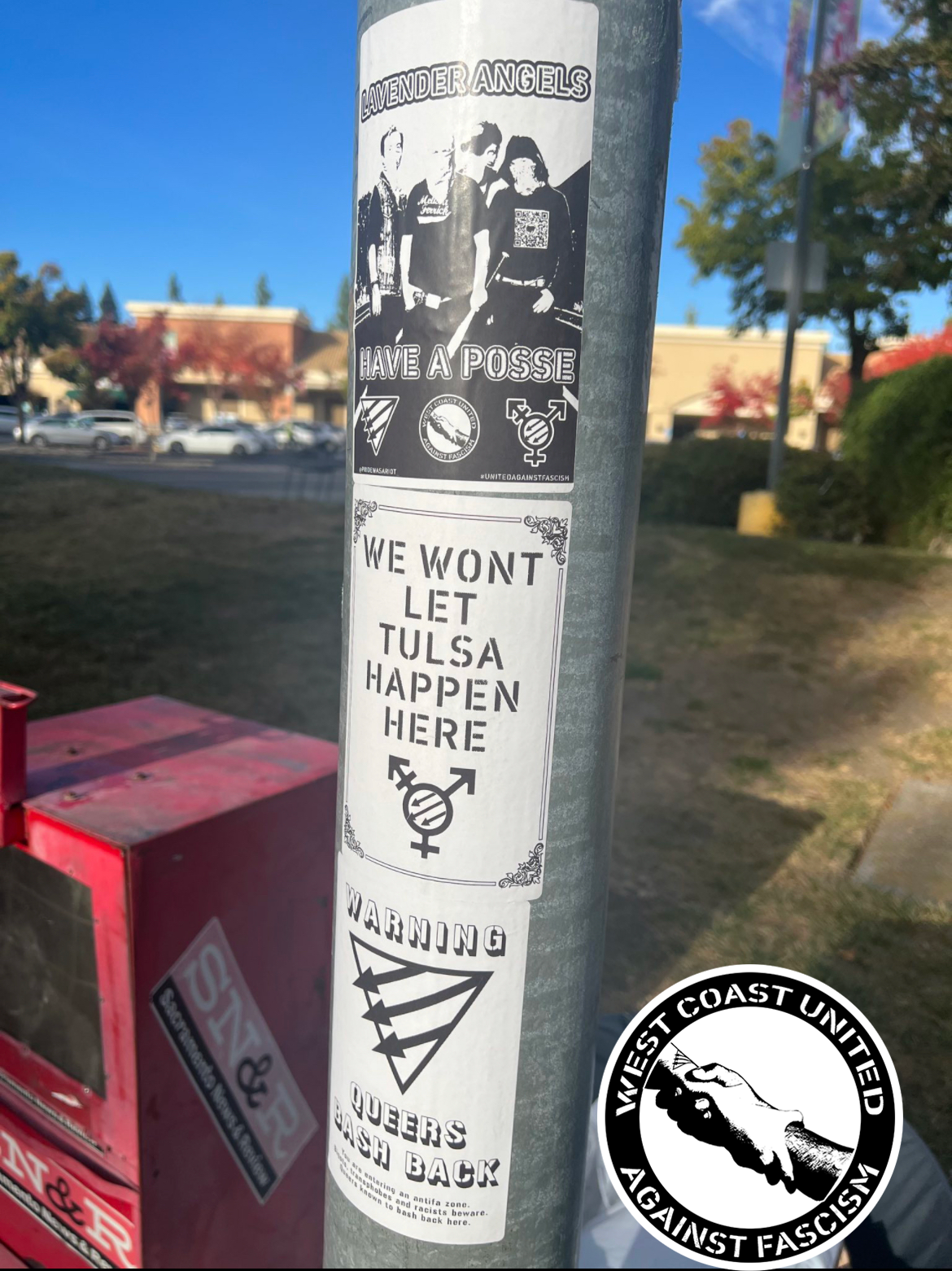 A metal post has three flyers adhered to it top to bottom. The background appears to be an outlet mall with cars. The first flyer reads "Lavender Angels Have A Posse." with the WCUAF logo, a Queer Front, and a Trans Front symbol. The second image reads "We Wont Let Tulsa Happen Here." with a Trans Front symbol. The third reads "Warning, Queers Bash Back" with a Queer Front symbol.