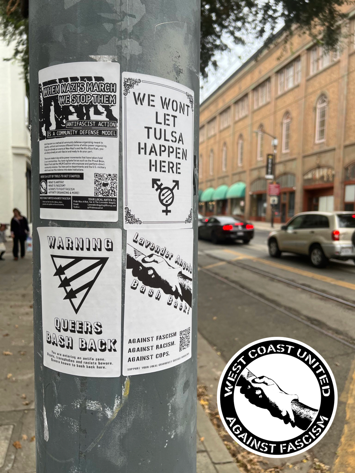 A metal post has four flyers adheared to it. The background appears to be city traffic with tall buildings surrounding it. Flyers are posted two by two. Top left reads "When Nazis March We Stop Them. Antifascist Action Is A Community Defense Model." with various text. Bottom Left reads "Warning, Queers Bash Back." with a Queer Front symbol across it. Top right reads, "We Wont Let Tulsa Happen Here." with a Trans Front symbol on it. Bottom right has two people locking harms which reads "Lavender Angels Bash Back. Against Fascism, Against Racism, Against Cops."