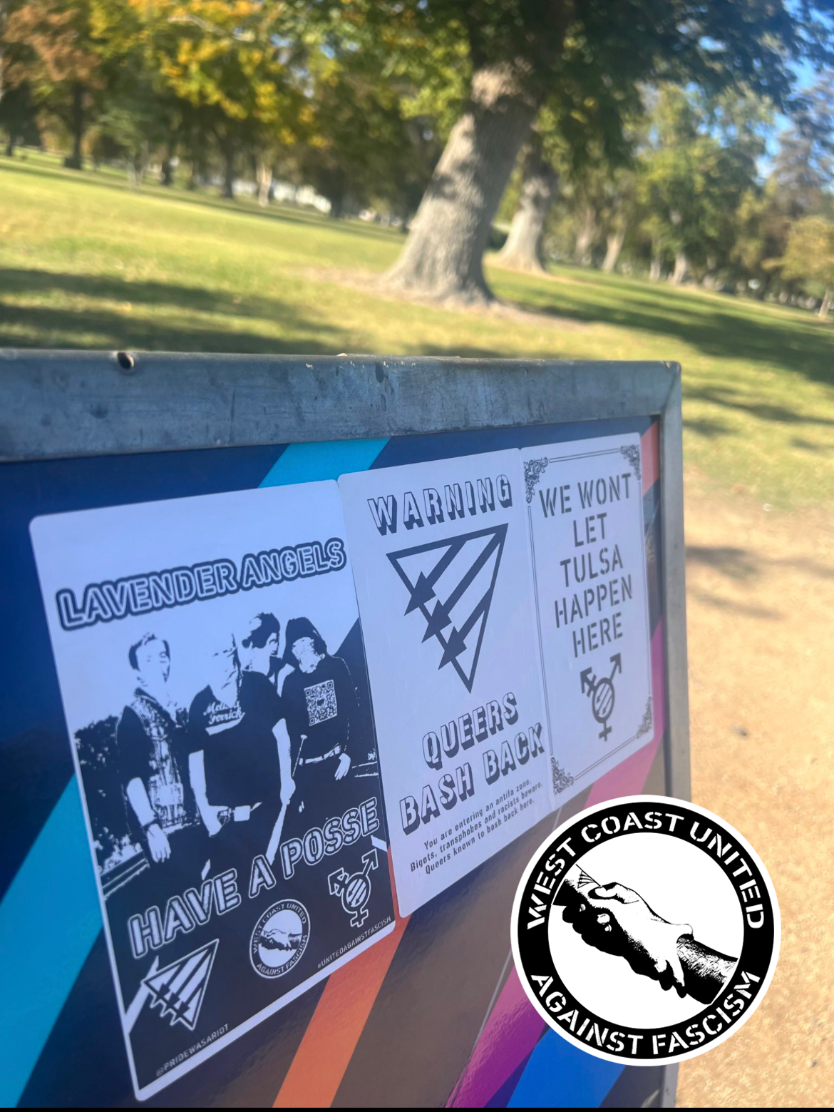 What appears to be the corner of a bus stop with a field of grass and trees behind it has three flyers adheared to the corner. From left to right, the first flyer reads "Lavender Angels Have A Posse." The image has a group of people standing, one carrying a bat. The bottom of it has the symbols for the Queer Front, WCUAF, and the Trans Front. The second flyer reads "Warning, Queers Bash Back." with the Queer Front symbol. The third reads "We wont let Tulsa happen here." with a Trans Front symbol across it.