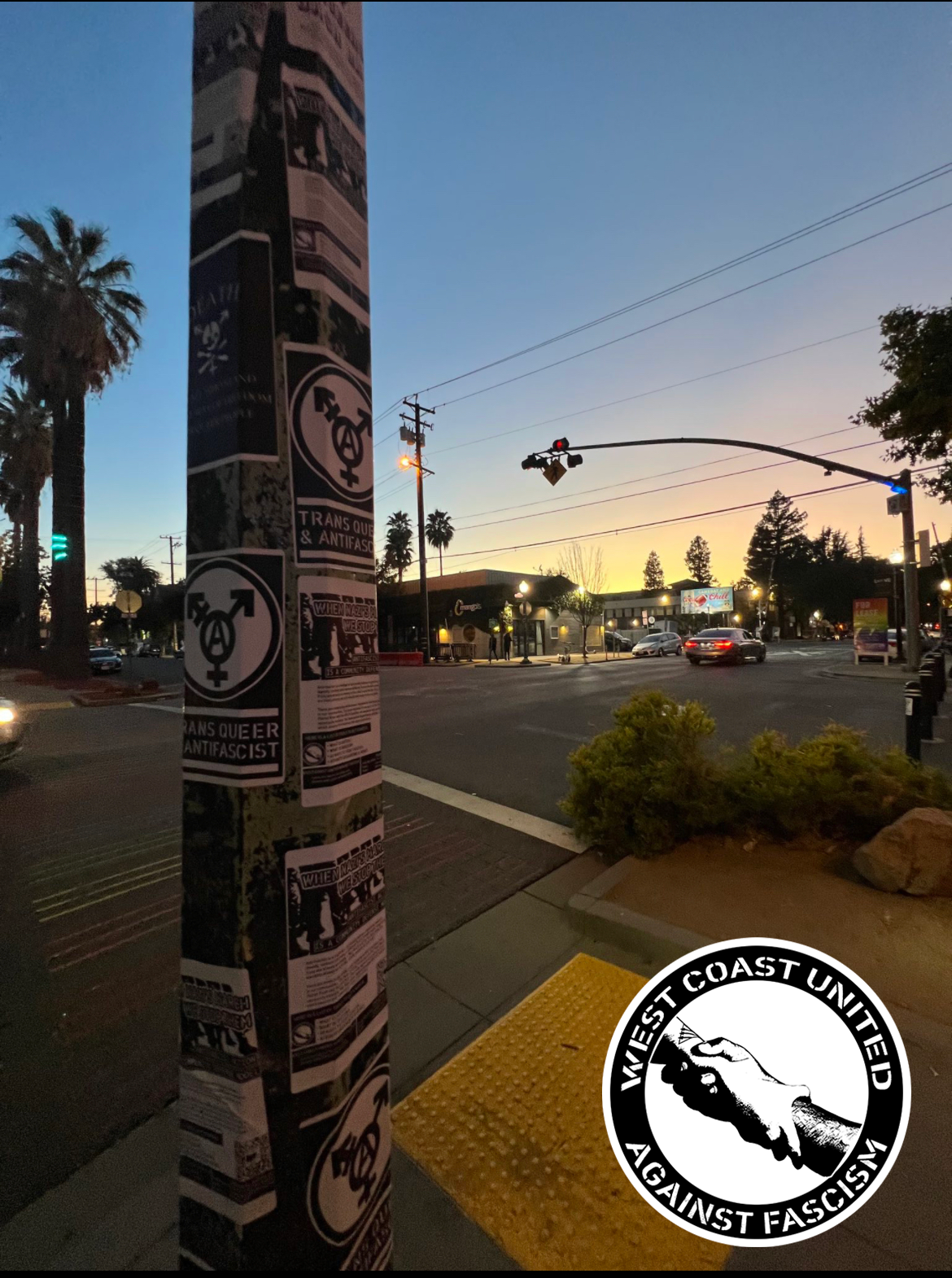 A lamp post sits in the foreground of a city intersection with a setting sun barely glowing in the distance. The metal post is covered in stickers, ranging from a Queer Mahkno, "Trans Queer and Antifascist" with a Transgender Anarchist logo, and flyers issued by West Coast United Against Fascism that has information how to begin antifascist work.