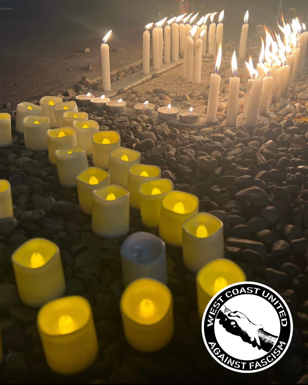 A row of battery powered candles and fire lit candles sit across a row of strewn pebbles and rocks across the ground. The darkness of the image is contrasted by the light from the candles.