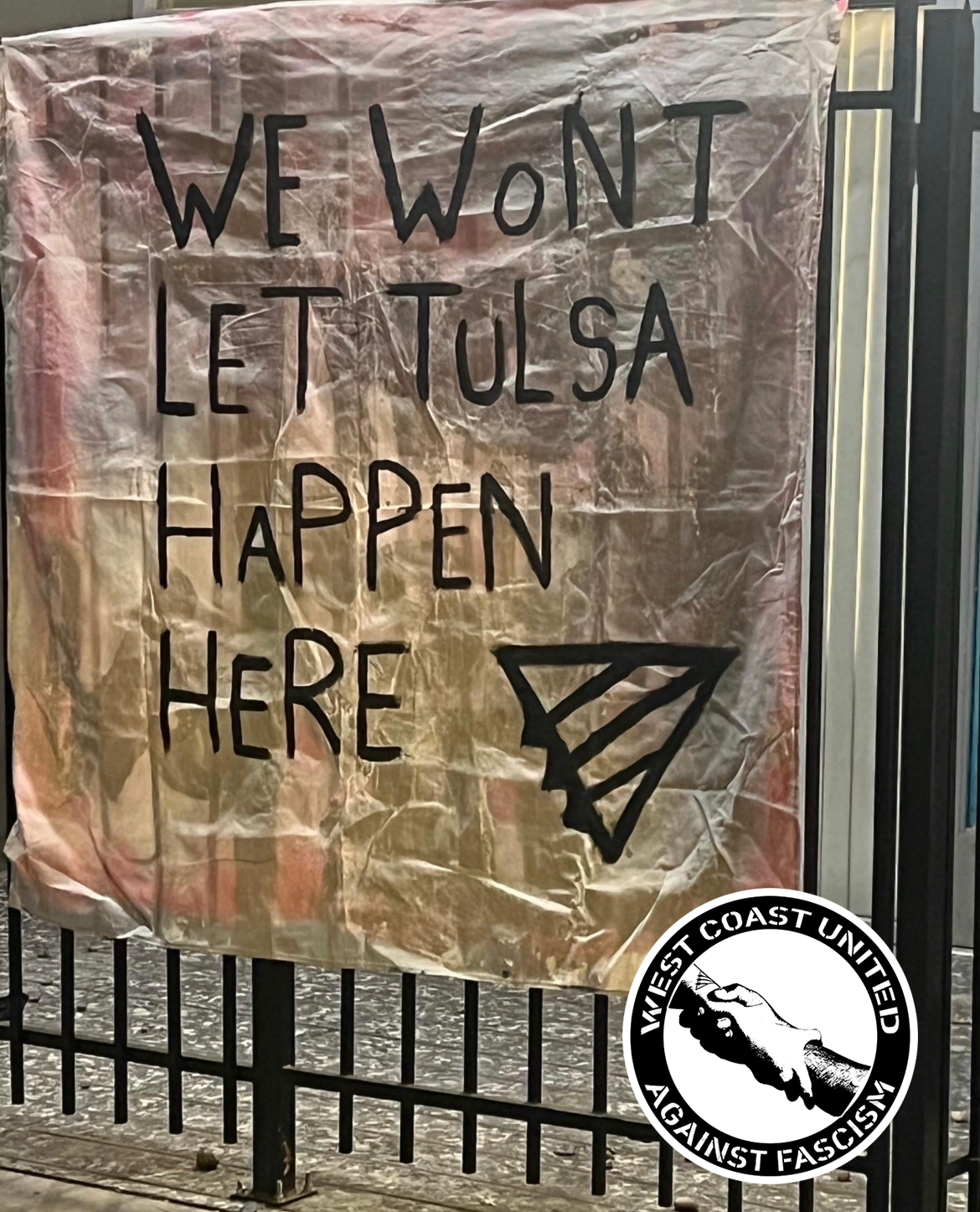 A metal fence line has a banner hanging from it that reads "We wont let Tulsa Happen Here." with a queer front painted on.