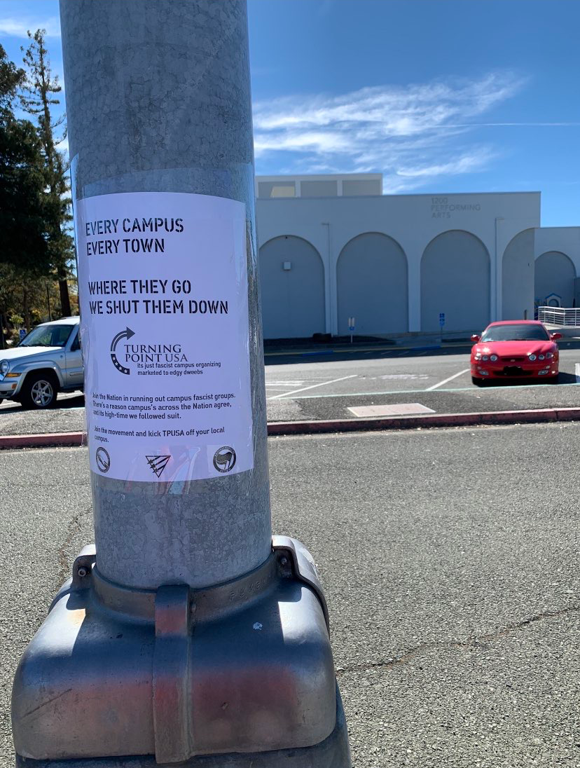 A metal post in a campus parkinglot has a flyer adhered to it. The lot has a few cars and buildings in the background. The flyer reads "Every Campus, Every Town, Where They Go We Shut Them Down" with the Turning Point USA symbol on it. Various text is difficult to read below it. The West Cost United Against Fascism symbol sits at the bottom next to a Queer Front symbol and an Antifascist Action symbol.