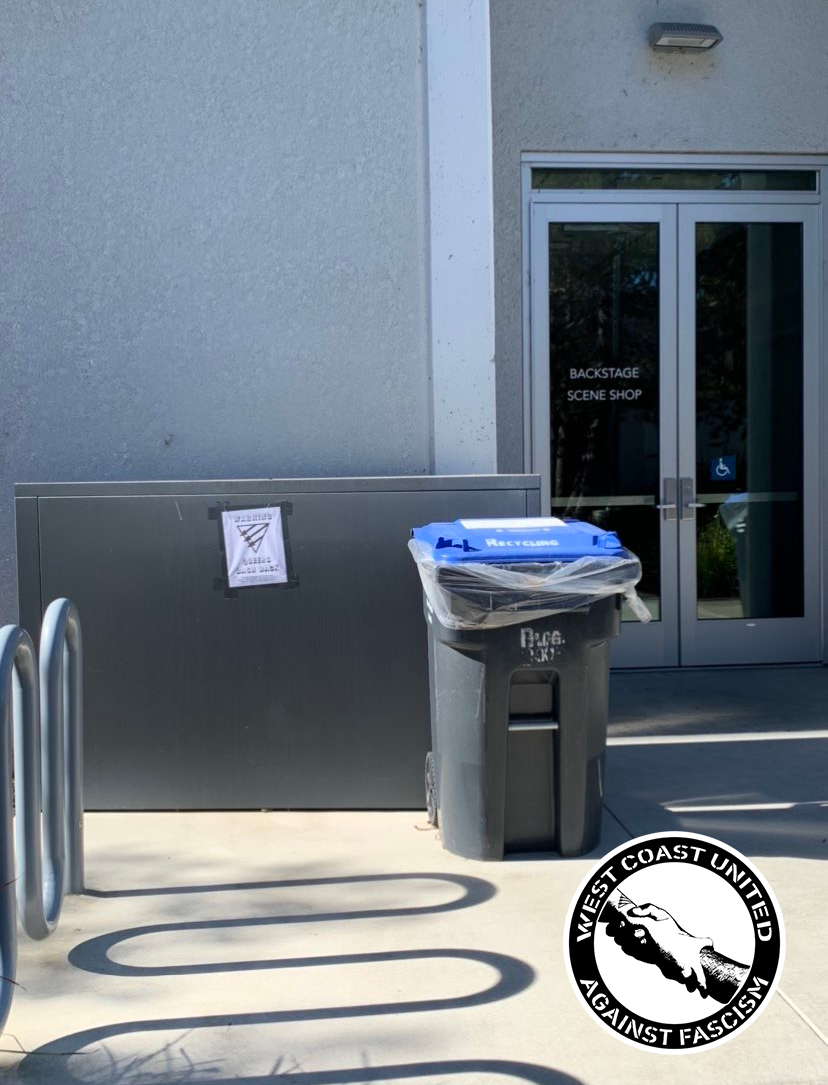 The entrance to a campus building is two glass doors. Near the entry way is a trash can and a bike rack. Against the wall to the entryway is a flyer adheared to the wall. It reads "Warning Queers Bash Back" with a queer front symbol on it.