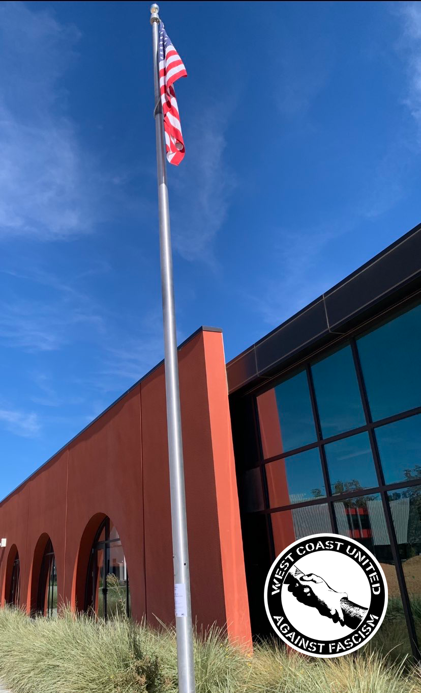 A flag pole with an american flag sits in front of a lage building with tall grass. Near the bottom appears to be literature flyered to it.