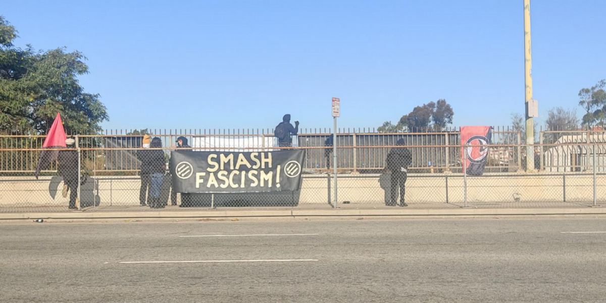 Antifascists in black wave flags and are surrounded by banners on a freeway overpass. One banner turned towards the camera reads "smash fascism!" with three arrows on the left and right of the text.