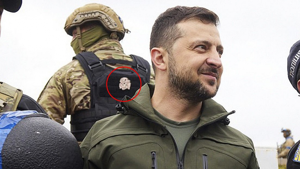 President Volodymyr Zelensky of Ukraine standing in front of Ukrainian soldiers wearing Neo-Nazi emblems on their plate carriers. Zelenksy is in a green jacket with a flared collor, with an overgrown chinstrap beard and mustache. His head is trim on the sides and with short, brown hair on top. The soldier behind him is in tan camo with a black plate carrier. The background is a grey, overcast sky.